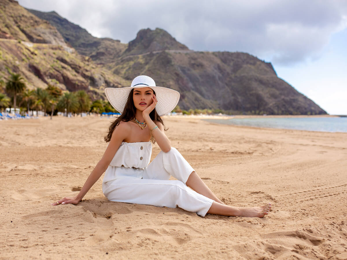 Modelka siedzi na piasku na plaży i prezentuje modne ubrania na wakacje. W tle morze, wzgórze i miasteczko.